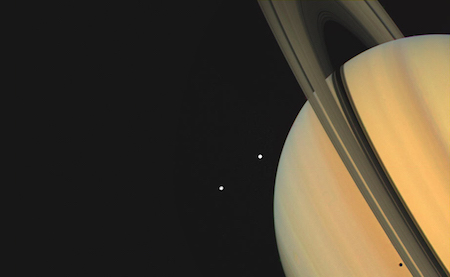 Một bức ảnh chụp Sao Thổ với các vành đai của nó ở một góc hướng lên trên. Bên cạnh sao Thổ là hai chấm trắng, là mặt trăng.