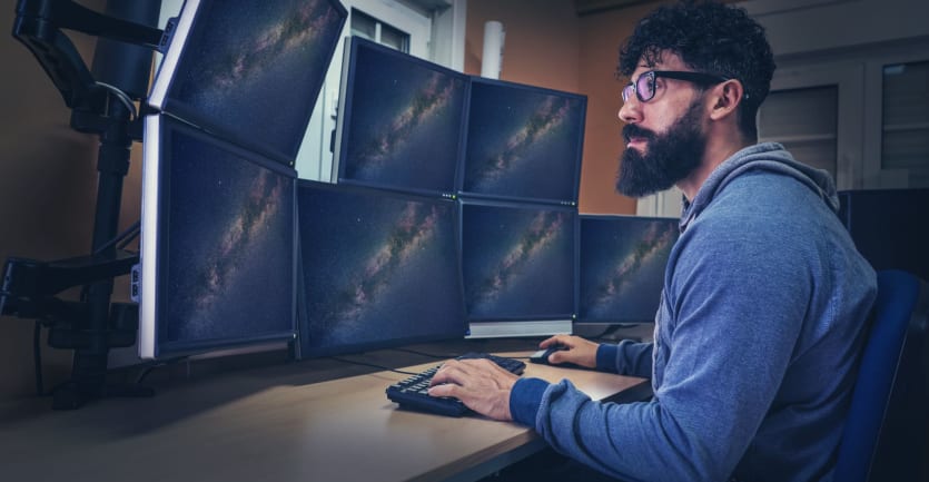 Một người đàn ông để râu đeo kính và mặc áo khoác trùm đầu ngồi trước một loạt màn hình máy tính hiển thị các bức ảnh về các hiện tượng thiên văn.